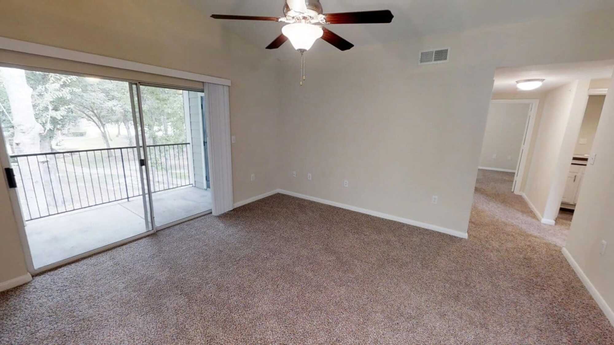 Empty carpeted room with beige walls, ceiling fan, and sliding glass door leading to a screened balcony.