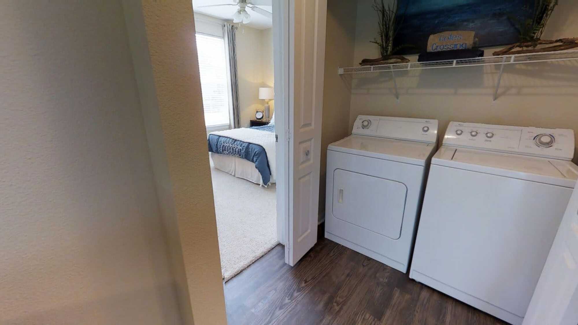 Laundry area with washer and dryer next to a carpeted bedroom with a bed and window, separated by a door.