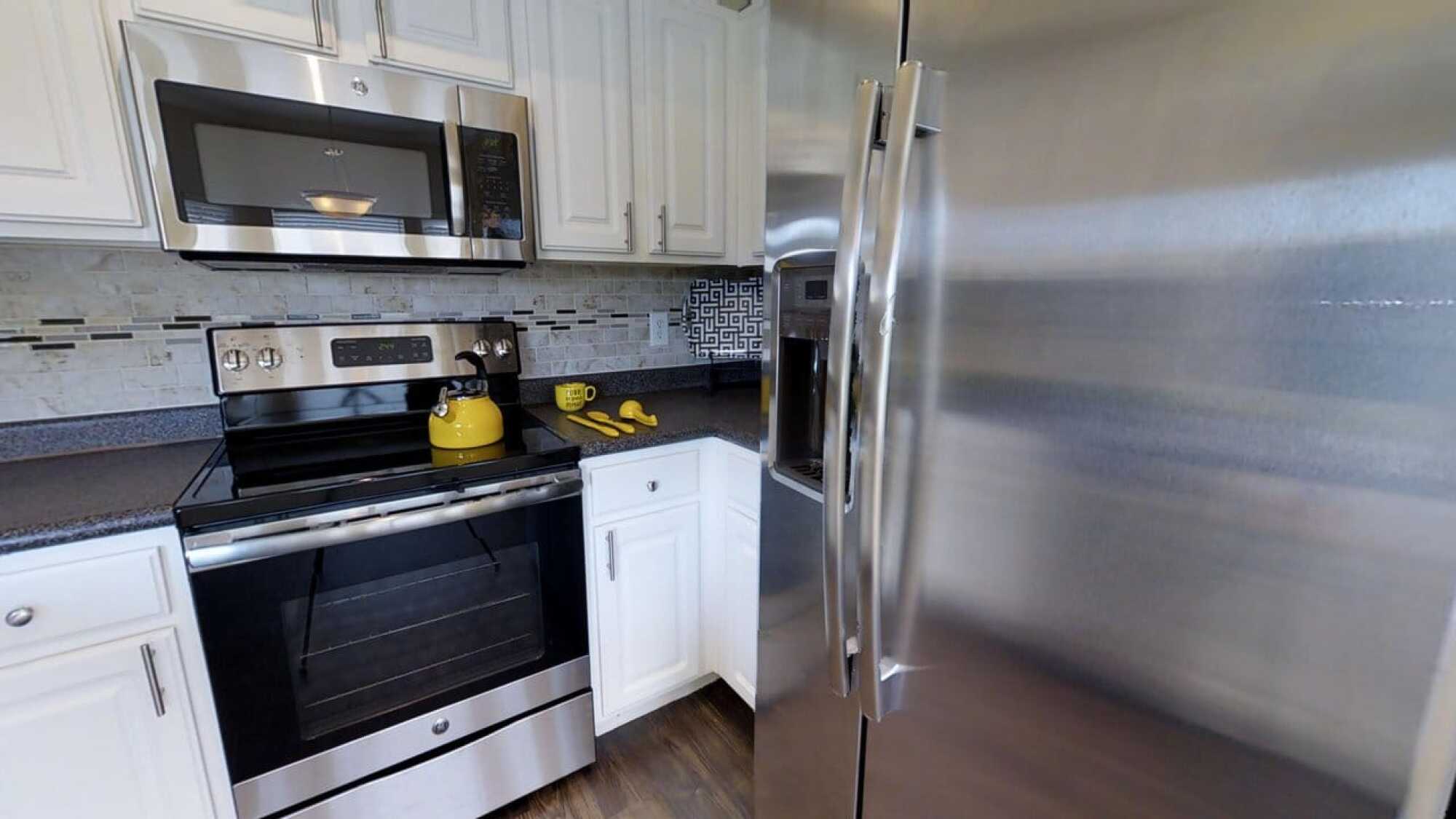 Modern kitchen with stainless steel appliances, white cabinets, yellow kettle, and bananas on a dark countertop.