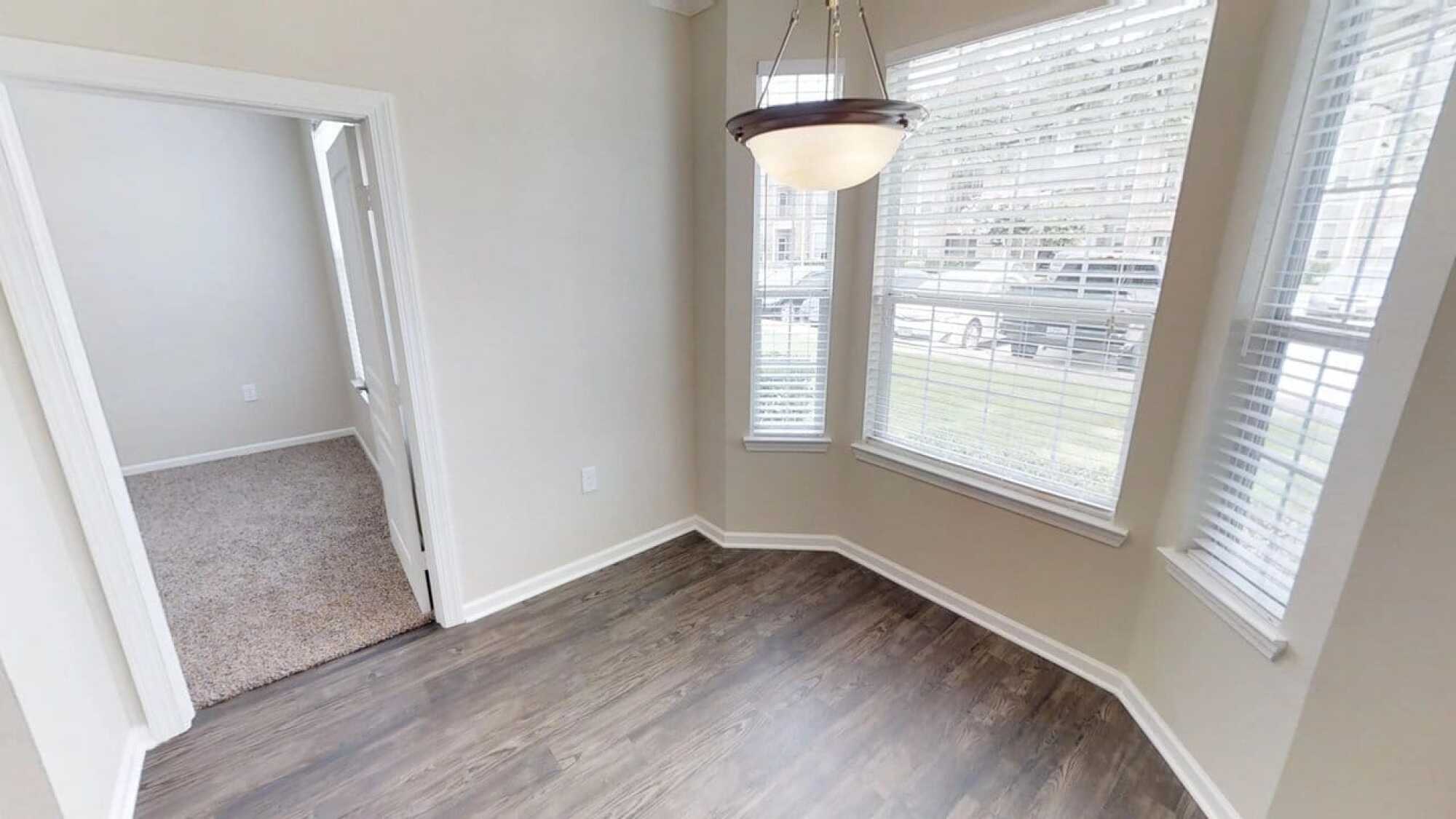 Empty room with wood floor, beige walls, three windows, and a ceiling light fixture hanging near the corner.