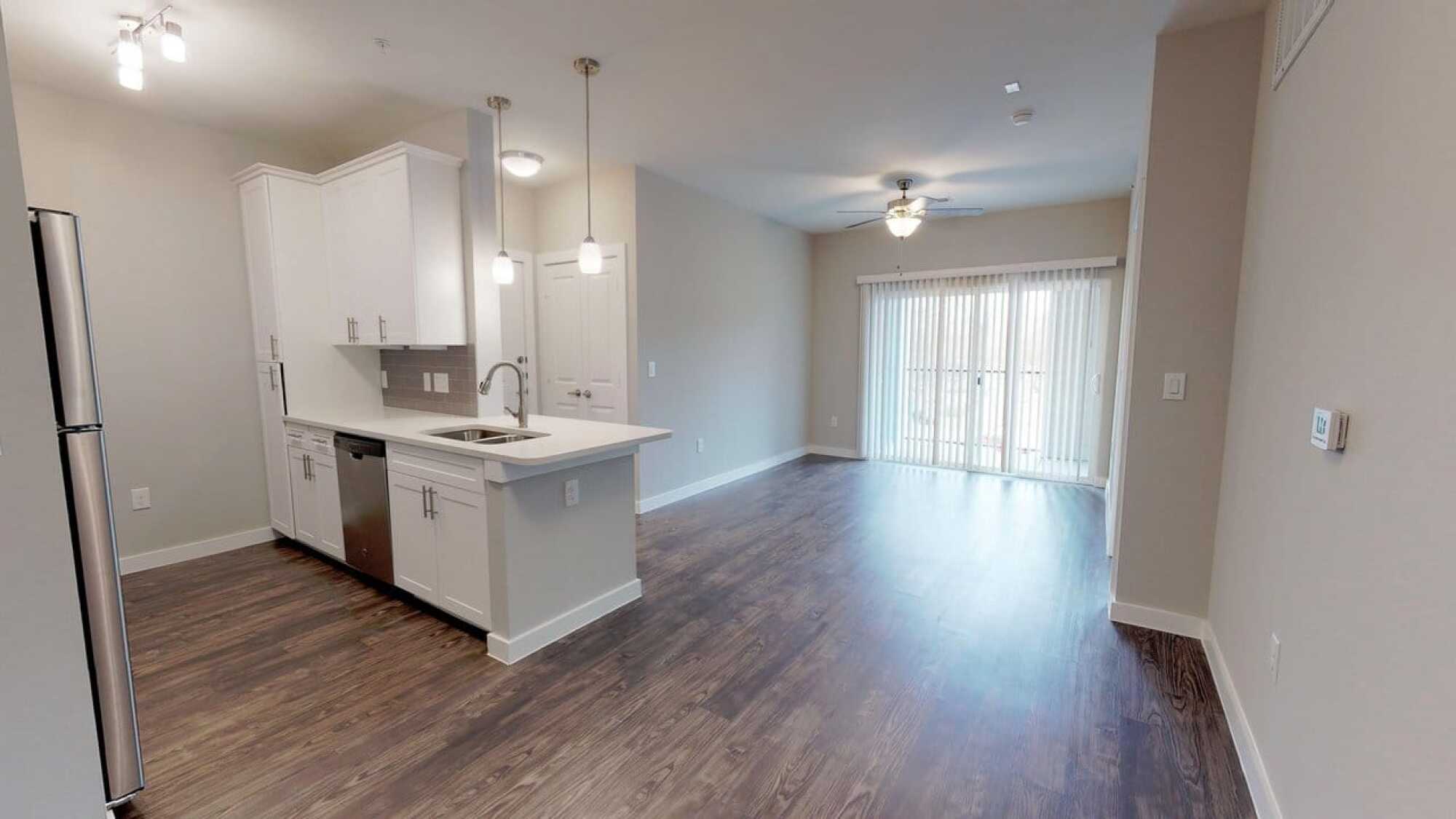 Modern apartment with white kitchen, pendant lights, dark wood floors, and sliding glass doors leading to a balcony.