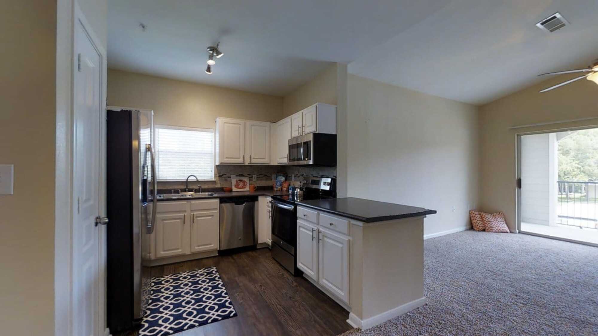 Modern kitchen with white cabinets, stainless steel appliances, and dark countertops, next to a carpeted living area with a sliding glass door.