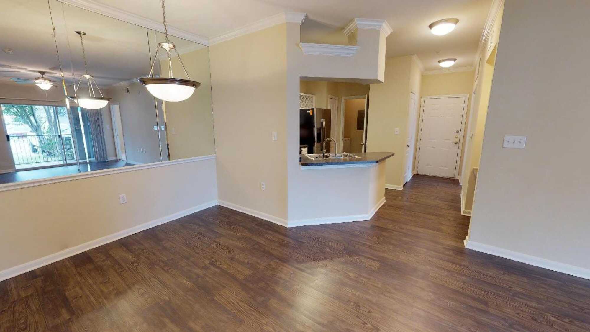 Empty apartment with wood floors, beige walls, a kitchen with stainless steel appliances, and a hanging light fixture.