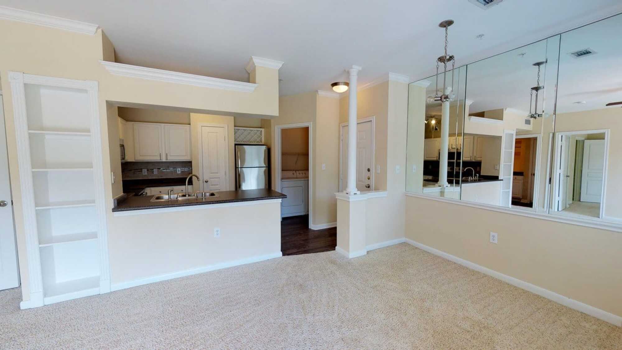 Open-plan living area with beige carpet, kitchen with white cabinets, and stainless steel appliances, plus mirrored wall accents.