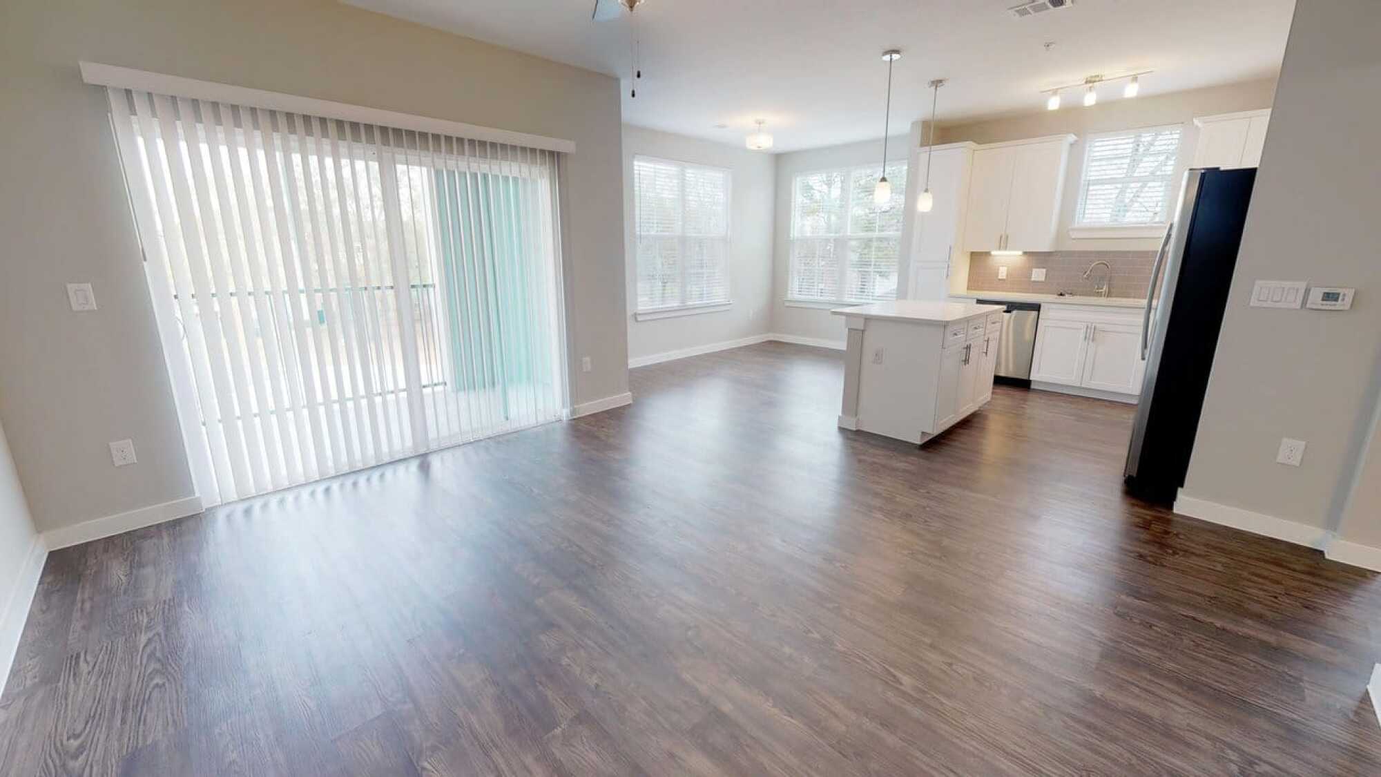 Bright, empty kitchen and dining area with wood floors, white cabinets, large windows, and glass sliding doors.