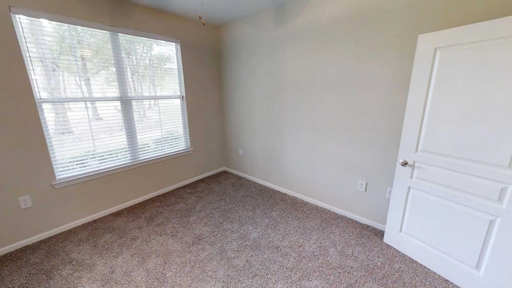 Empty carpeted room with beige walls, a window with blinds, and natural light coming in from outside.