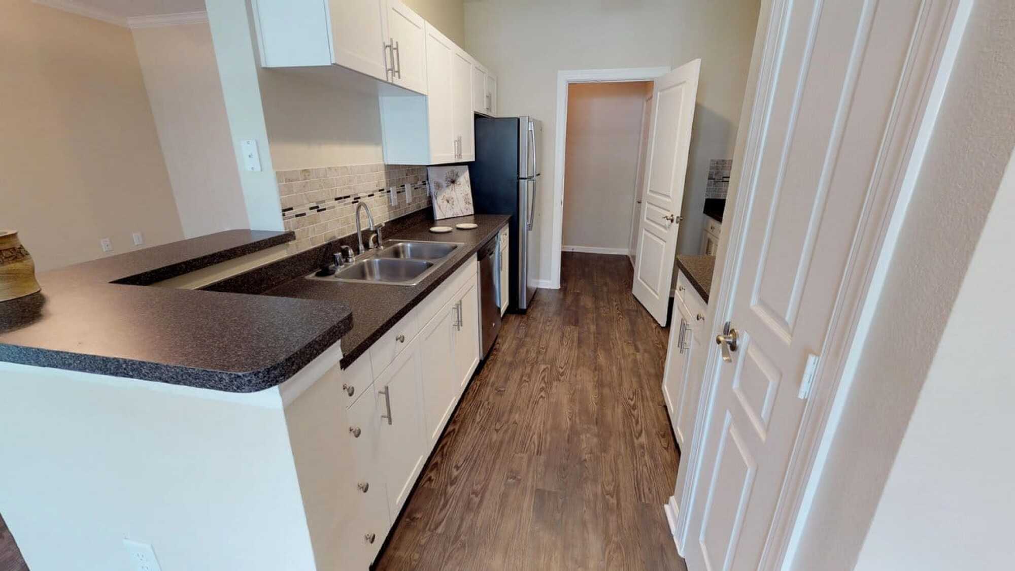 Modern galley kitchen with white cabinets, dark countertops, stainless steel appliances, and wood-look flooring.