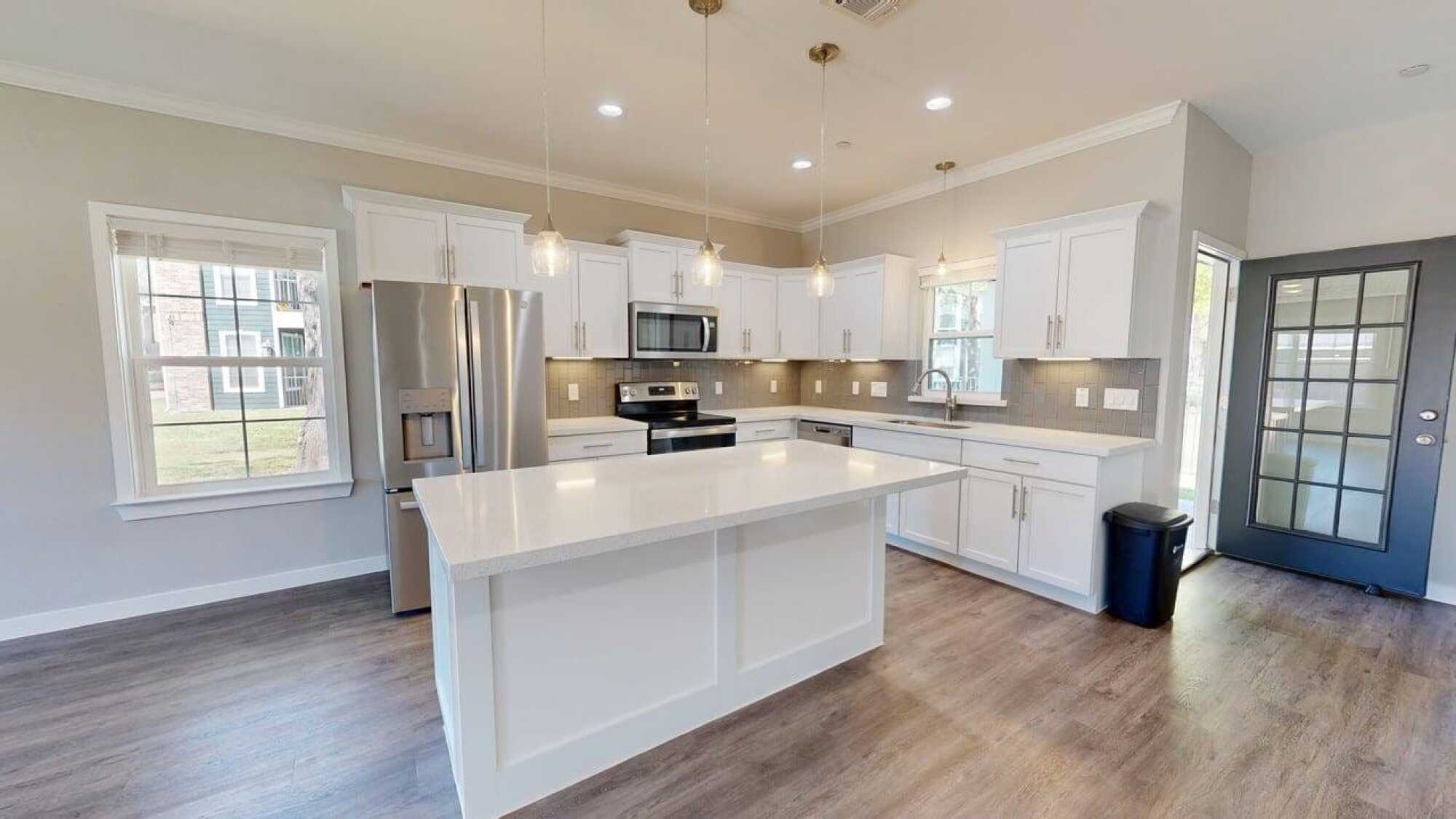 Modern kitchen with white cabinets, stainless steel appliances, pendant lights, and a large island on wood flooring.