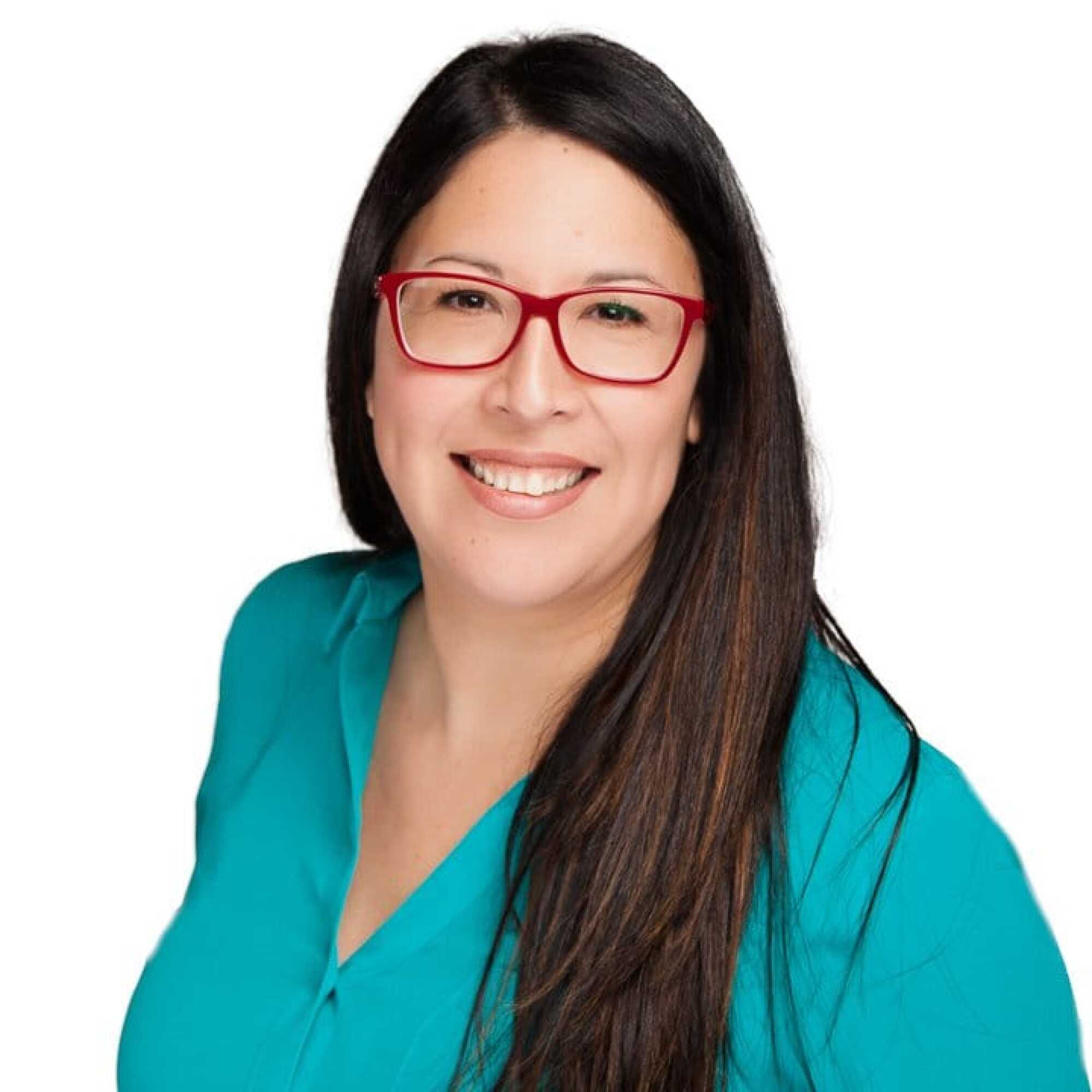 Woman with long dark hair, red glasses, and a teal top smiling against a plain white background.
